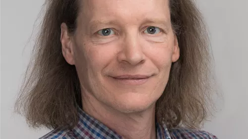 Portraitfoto von Christopher Coenen. Er trägt ein kariertes Hemd und posiert vor einem grauen Hintergrund. Seine Haare sind schulterlang