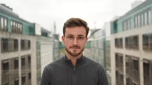 Portraitfoto von Maurice Höfgen. Er trägt kurze Haare, Bart, einen Pullover und Brille. Er schaut in die Kamera. Im Hintergrund sind auf beiden Seiten Gebäude zu sehen.