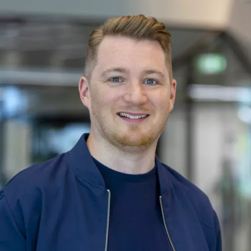 Eine Person steht in einem modernen Innenraum mit unscharfem Hintergrund. Die Person trägt eine dunkelblaue Bomberjacke mit einem silbernen Reißverschluss sowie ein dunkelblaues T‑Shirt. Das Haar ist kurz an den Seiten und länger nach hinten gestylt. Hinter der Person sind Glasflächen und metallische Strukturelemente eines Gebäudes zu erkennen, die auf eine Büro- oder Empfangssituation hindeuten. Die Beleuchtung wirkt hell und gleichmäßig.