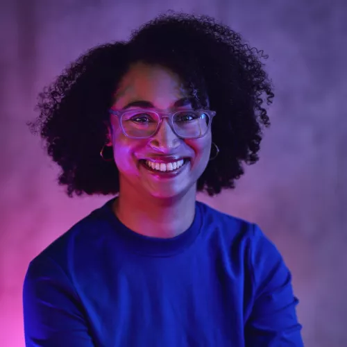 Eine Person mit offenen Locken und Brille strahlt in die Kamera. Sie trägt ein dunkelblaues Oberteil und wird mit rosa Licht indirekt angestrahlt.
