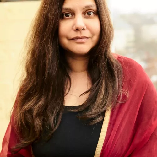 Auf dem Portrait-Bild ist Mithu Sanyal mit langen braunen Haaren zu sehen und trägt eine rote Jacke. Sie schaut freundlich ohne zu lachen in die Kamera.