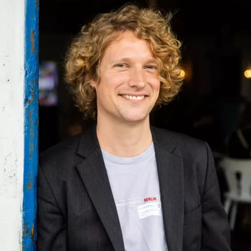 Ein Portraitfoto von Niklas Kossow, einem Mann mitte 30 mit blonden Locken, er trägt ein Lila TShirt und ein Sakko.