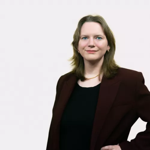 Porträt von Konstanze Renken mit schulterlangen dunkelblonden Haaren und blauen Augen. Sie trägt einen bordeauxroten Blazer über schwarzem Top und eine goldene Halskette. Pose mit Hand in der Hüfte vor neutralem hellgrauem Hintergrund, sie blickt freundlich mit geschlossenem Mund in die Kamera.