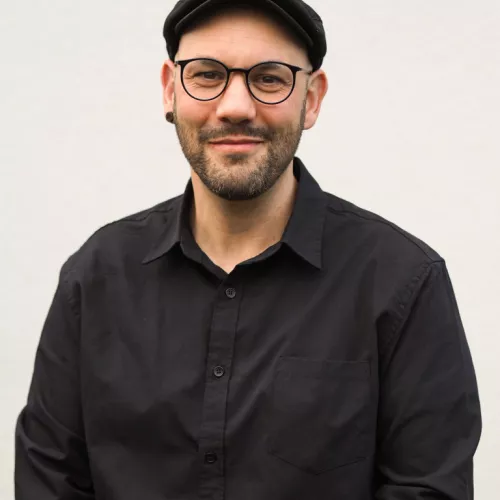 Portraitfoto mit beigem Hintergrund. Schwarzes Hemd, schwarze Schiebermütze.