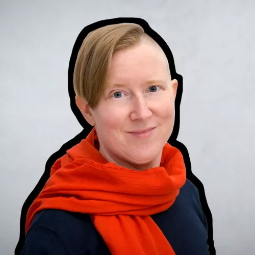 Annika Brinkmann trägt einen Orangen Schal und einen dunkelblauen Pullover. Sie lächelt in die Kamera. Ihre rötlichlichen Haare trägt sie an den Seiten kurz, die längeren Haare fallen ihr über das rechte Ohr.