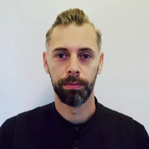 Profilfoto bis zu den Schultern. Kameraperspektive von vorn. Weißgrauer Hintergrund. Im Vordergrund ein Typ Mitte dreißig mit schwarzen Hemd, Bart, und langen Haaren nach hinten gekämmt. 