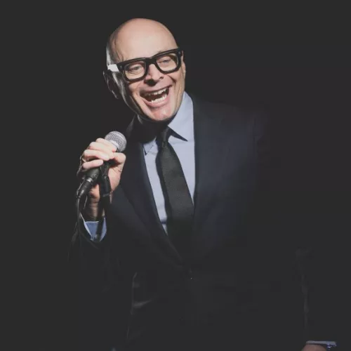 A bald white man in a suit and tie hold a microphone and is laughing loudly.