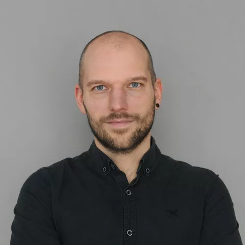 Männliche Person mit sehr kurzen Haaren, Bart und dunklem Hemd.