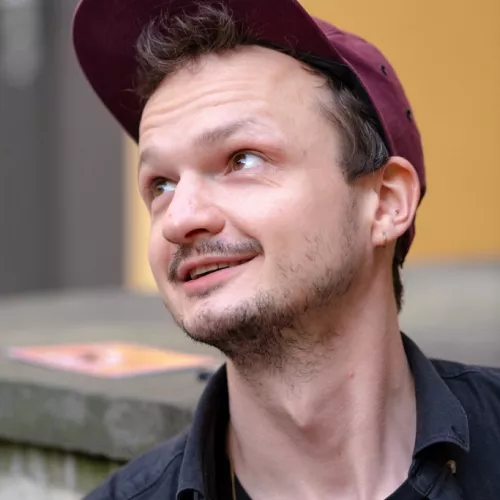 Das Bild zeigt Fabians Kopf, sein Blick geht nach links oben. Er trägt eine weinrote Schirmmütze und ein schwarzes Hemd, darunter ein schwarzes T-Shirt. Er lächelt in die Richtung, in die er schaut.