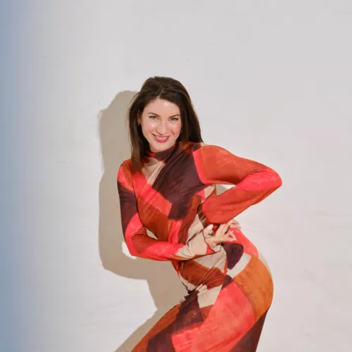 Porträt einer Person in orangefarbenem Kleid vor einer weißen Wand. Am linken Rand ich ein feiner blauer Streifen.