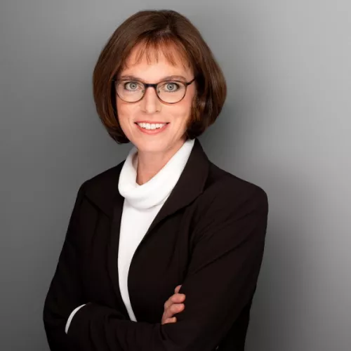 Lächelnde Frau mit Brille und schwarzem Blazer sowie weißem Pullover steht vor neutralem Hintergrund.
