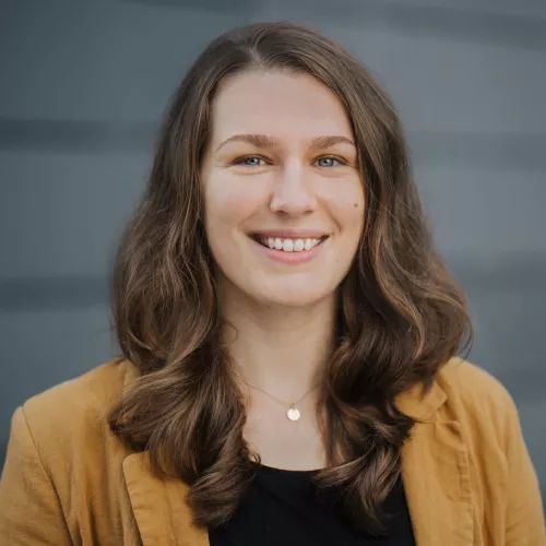 Marlen Förderer lächelnden vor grauem Hintergrund mit schulterlangem braunem Haar, gelbem Blazer und schwarzem Oberteil .