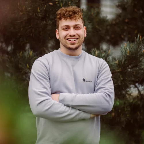 Auf dem Foto ist ein junger Mann zu sehen. Er hat rot-braune, kurze, lockige Haare, trägt einen fliederfarbenen Pullover vom Abenteuer Regenwald e.V. und lächelt fröhlich in die Kamera. Seine Arme sind vor dem Überkörper verschränkt. Im Hintergrund befindet sich ein Baum.