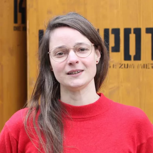 Frau in rotem Pulli mit Brille und offenen Haaren vor Holzobjekten