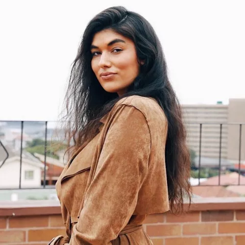 Photo of woman with long black hair and tanned skin, wearing a beige coat. 