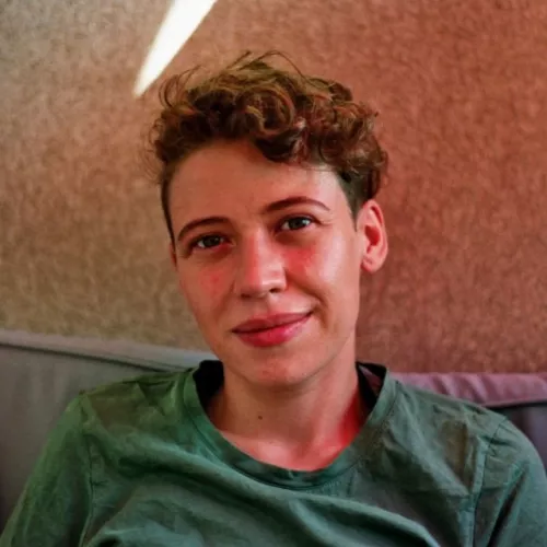 Das Bild zeigt ein frontales Portraitfoto von Cin. Cin guckt entspannt nach vorne, der Kopf ist leicht nach links geneigt. Cin hat kurze, braune Locken, volle Lippen, leichte Lachfältchen um die Augen und ist weiß. Cin trägt ein Tshirt in einem mittleren Grünton. Im Hintergrund ist eine beige Hauswand und ein graues Polster an die Cin angelehnt ist. Ein Sonnenstrahl bricht durch den oberen Bildrand links an Cins Kopf vorbei, das Bild hat einen leicht orange-roten Einschlag und wirkt sommerlich.