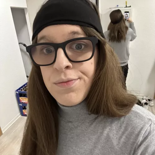 Eine junge Frau lächelt für ein Selfie in die Kamera. Sie trägt einen grauen Rollkragenpullover und glatte, braune Haare unter einem schwarzen Stirnband. Im Hintergrund spiegelt sich ihr Rücken durch einen großen Wandspiegel.
