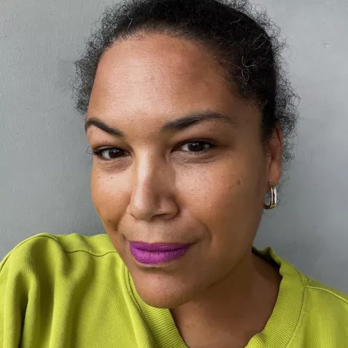 Porträtfoto von Olivia [Nachname]. Sie blickt leicht lächelnd in die Kamera, trägt einen gelbgrünen Pullover und lila Lippenstift. Hintergrund ist eine graue Wand