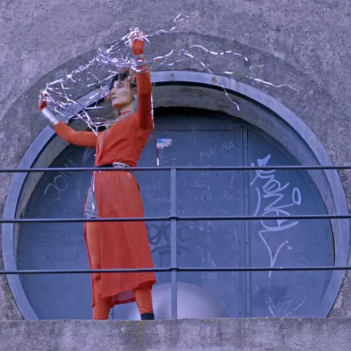 A portrait of Jessika Khazrik standing next to a silver sphere against a concrete backdrop with a circular gate, holding with her raised arms a web of scintillating threads