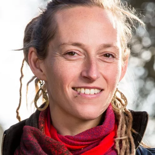 Das Bild zeigt eine Person mit einem freundlichen Lächeln. Sie hat helle Haut, trägt Dreadlocks und auffällige goldene Ohrringe. Ein roter Schal und eine dunkle Jacke ergänzen ihr Erscheinungsbild. Der Hintergrund ist unscharf, was auf eine Aufnahme im Freien bei Tageslicht hinweist.