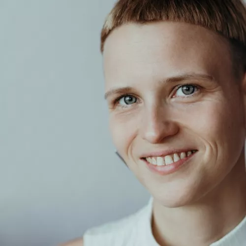 Manuela hat kurze blonde Haare, dreieckige Ohrringe und lächelt in die Kamera.