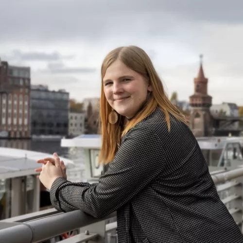Eine Frau mit langen Blonden Haaren steht an ein Geländer gelehnt vor der Spree und schaut in die Kamera. Im Hintergrund ist die Oberbaumbrücke zu sehen. 