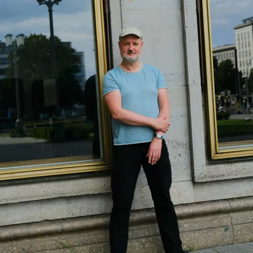 Das Bild zeigt den Journalisten Martin Kaluza. Er trägt eine Mütze und steht vor der großen Fensterscheibe eines Gebäudes.