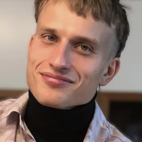 Das Bild zeigt als Portraitaufnahme eine männlich gelesene Person in Hemd über schwarzen Rollkragenpullover. Halbkurze dunkelblonde Haare, blaue Augen.