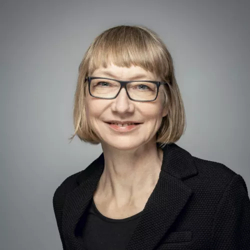 Porträtfoto von Sabine Pfeiffer vor grauem Hintergrund. Sie lächelt in die Kamera, hat kinnlange blonde Haare und trägt eine schwarze Brille. Bekleidet ist sie mit einem schwarzen Blazer über einem schwarzen T-Shirt.