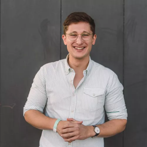 Das Foto zeigt einen jungen Mann mit braunen Haaren und Brille in einem hellen, gestreiften Hemd vor grauer Wand. Er lächelt in die Kamera.