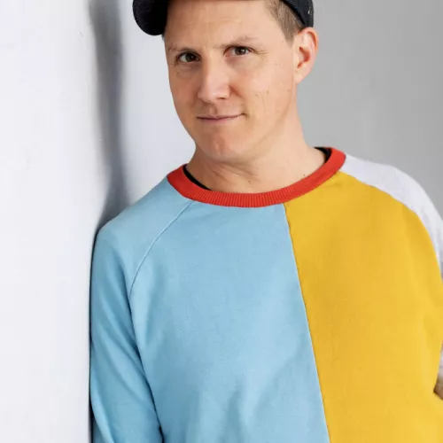 Foto des Autoren Sebastian 23. Er trägt einen bunten Pullover und eine Schiebermütze und lehnt lächelnd an einer weißen Wand.