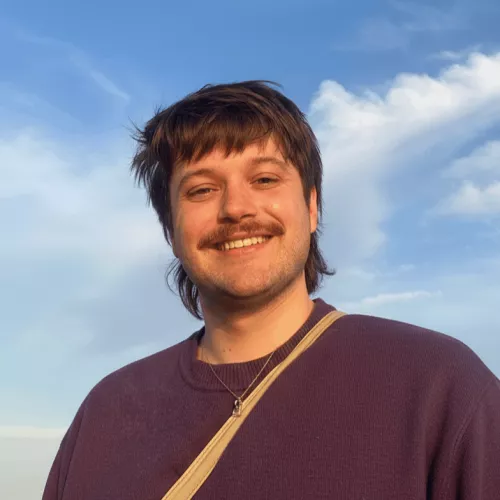 Das Bild zeigt das Profilfoto von Lucas Braun. Der Mitt-Zwanziger mit braunen Haaren und Schnauzbart lächelt in die Kamera vor einem leicht bewölkten Himmel.