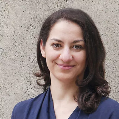 Porträtfoto einer Frau mit schulterlangem, dunklem Haar. Sie blickt frontal in die Kamera und lächelt leicht. Sie trägt ein dunkelblaues Oberteil. Der Hintergrund ist eine helle, strukturierte Steinwand.