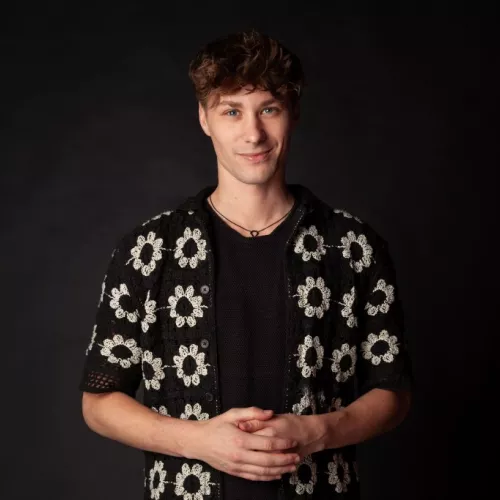 Simon steht vor einem schwarzen Hintergrund und trägt einen weiß-schwarzen Blumencardigan. 