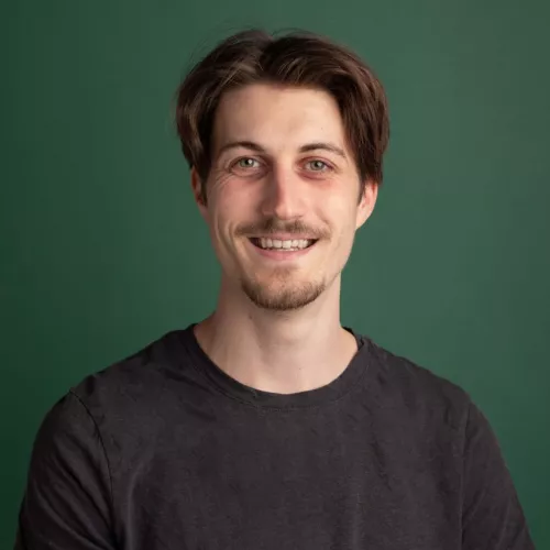 A man smiling in front of a green photo studio background. Brown hair and grey T-shirt.