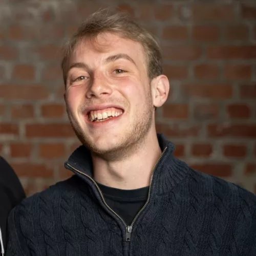 Stefan Wilhelm (ein blonder Mann Mitte 20) steht lächelnd vor einer Backsteinwand. Er trägt einen dunkelblauen Pullover.