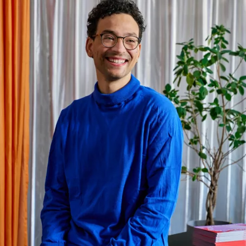 Quincey Stumptner von der Seite photographiert. Er lacht, trägt eine schwarze Brille und trägt einen blauen Pullover.