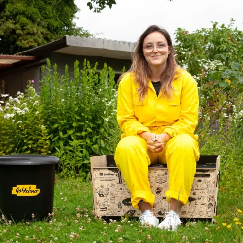 Tanja sitzt im gelben Overall auf einer Trockentoilette von Goldeimer und lächelt in die Kamera. Im Hintergrund sind blühende Sträucher.