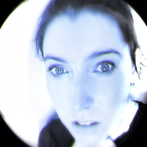 Photograph of the artist Lyndsey Walsh, cropped in a circular shape with a blue tint. Lyndsey has dark brown hair and pale white skin. The artist is looking directly at the camera.