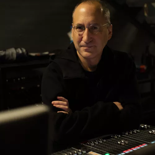 A white man in his mid 50s wearing thin framed glasses sits behind an audio mixing desk