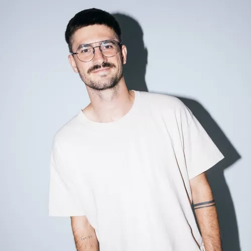 Auf dem Bild Alex Elsner zu sehen. Er trägt ein weißes T-Shirt, eine Brille it dünnem Gestell, hat braune Haare und braunen Bart.