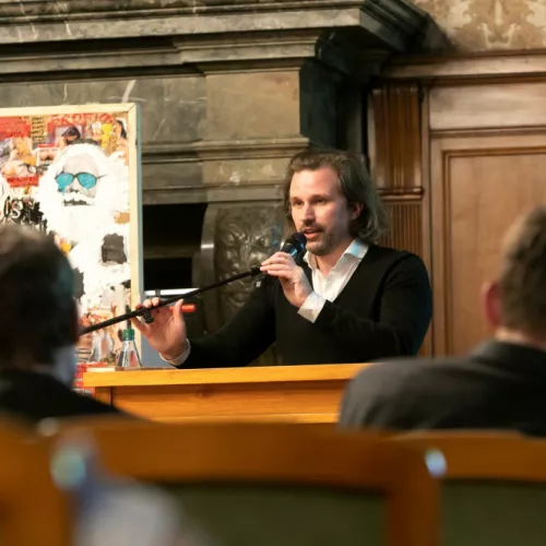 ein Mann mit längeren Haaren an einem Rednerpult, der Saal wirkt festlich