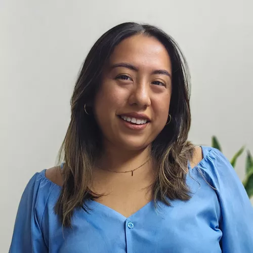 Smiling woman with shoulder-length dark hair wears a blue blouse and small hoop earrings, facing the camera against a neutral background.