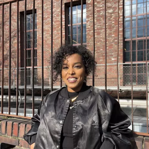 The image depicts Feven M. Keleta of SUPERRR Lab, in front of a brick building and fence. She is leaning against the fence and smiling towards the camera, wearing a black jacket, pants and shirt. 