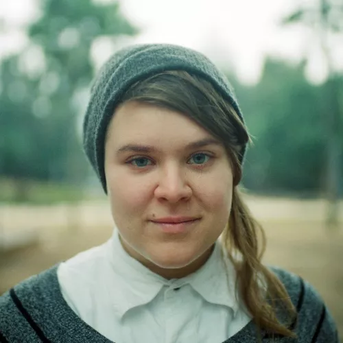 a blurred background of trees and tan ground; in front a person with brownish-blonde hair, a grey hat, light skin and blue eyes looks directly at the camera. they are wearing a white colloared shirt with a grey and black stripped jumper over that