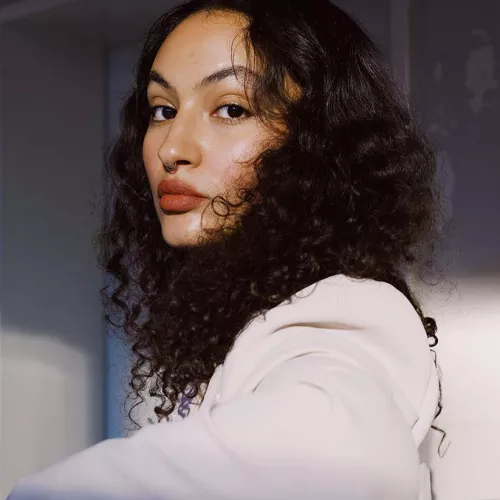 Anja trägt einen cremefarbenen Blazer und blickt mit angelegtem linken Arm über ihre linke Schulter direkt in die Kamera. Sie trägt ihr lockigen Haar offen und im Hintergrund ist ein weißer Türrahmen zu erkennen. 