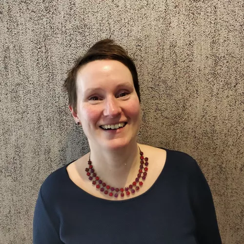 ein Foto von Nadja Ullrich. Sie hat braune kurze Haare und trägt ein petrolfarbenes Oberteil und eine rote Strasskette