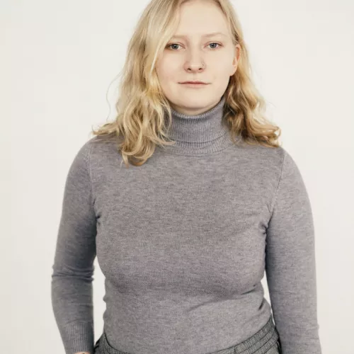 A young woman with shoulder-length blonde hair stands facing the camera against a plain light background. She wears a gray turtleneck sweater and gray plaid trousers, with one hand resting in her pocket. Her expression is neutral, and she has a small nose ring.